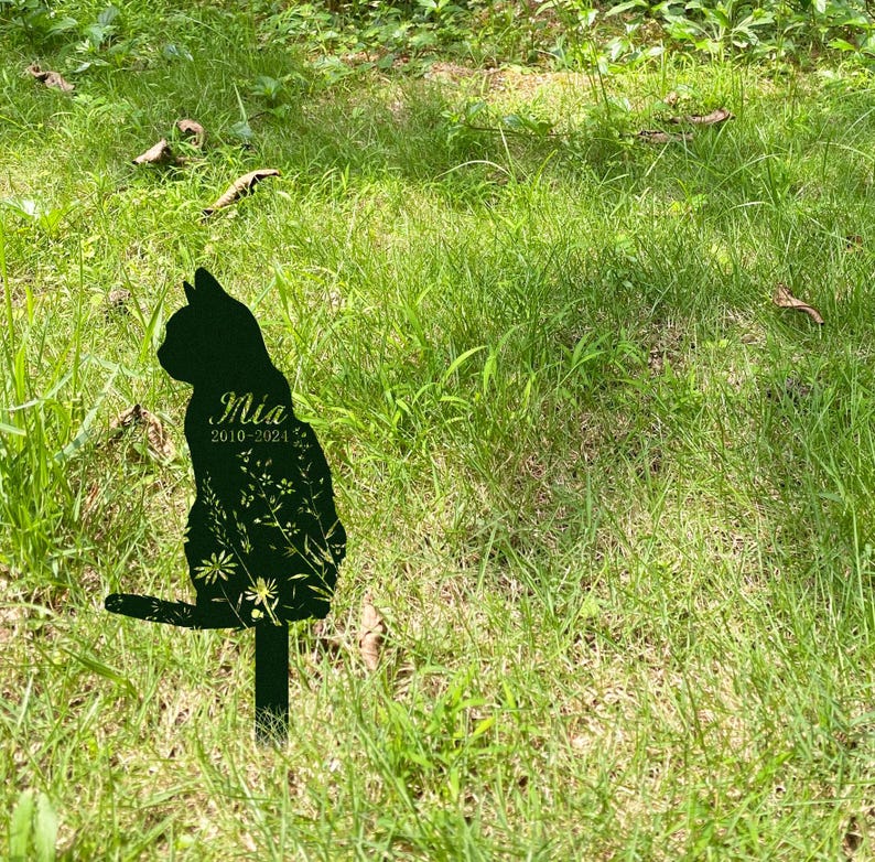 May include: A black metal cat silhouette garden stake with the name "Mia" and the years "2010-2024" etched on the front. The cat is decorated with floral designs and is placed in a grassy area.