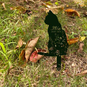 May include: A black metal cat silhouette garden stake with the name "Mia" and the years "2010-2024" cut out. The cat is sitting with its tail up, and the design includes floral cutouts. The stake is in a grassy area with fallen leaves.
