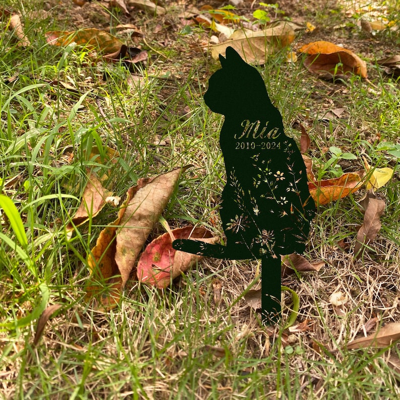 May include: A black metal cat silhouette garden stake with the name "Mia" and the years "2010-2024" cut out. The cat is sitting with its tail up, and the design includes floral cutouts. The stake is in a grassy area with fallen leaves.