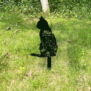 May include: A black metal cat silhouette garden stake with the name "Mia" and dates "2010-2024" in a cursive font. The cat's body is filled with a floral design. The stake is placed in a grassy area.