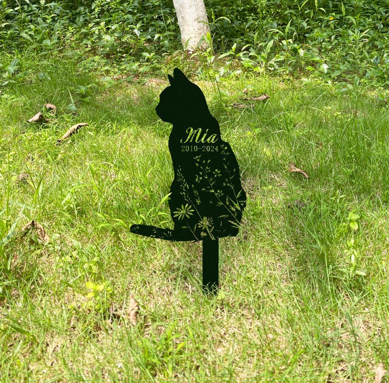 May include: A black metal cat silhouette garden stake with the name "Mia" and dates "2010-2024" in a cursive font. The cat's body is filled with a floral design. The stake is placed in a grassy area.