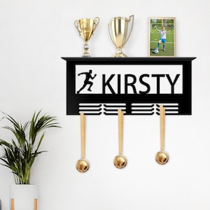 Puede incluir: Estante de pared de metal negro para trofeos con el nombre "Kirsty" y una silueta de un corredor. El estante tiene tres ganchos y un pequeño marco de fotos con una foto de un niño jugando al fútbol.