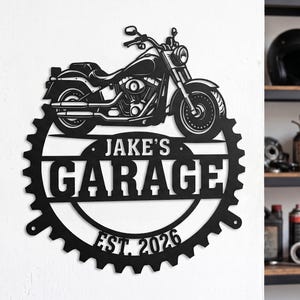 May include: A black metal wall sign featuring a motorcycle silhouette positioned above a gear-shaped frame. The text "JAKE'S GARAGE" is centered within the gear, with "EST. 2026" printed along the bottom edge of the circular metal structure.