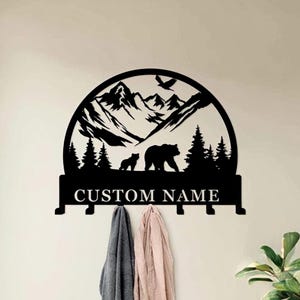 May include: Black metal wall art depicting a mountain scene with bears and trees. The design includes the text "CUSTOM NAME" and hooks for hanging items. Two garments hang from the hooks. A bird flies above the mountains.