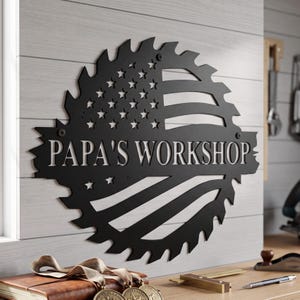 May include: A circular black metal wall sign shaped like a saw blade featuring a cutout American flag design and the text "PAPA'S WORKSHOP" in bold, capitalized letters across the center, secured with visible screws at the top and bottom.