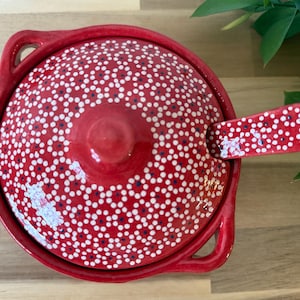 May include: Red ceramic serving dish with a white and blue polka dot pattern and a matching lid. The dish has two handles and a small spoon.