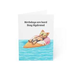 May include: A greeting card with a cartoon dog on an ice cream cone float in water. The dog has cucumber slices on its eyes. The card reads "Birthdays are hard, Stay Hydrated."