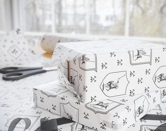 Może przedstawiać: Dwa białe pudełka prezentowe owinięte w czarno-biały papier ozdobny. Papier przedstawia powtarzający się wzór rycerza na koniu z tarczą i skrzyżowanymi mieczami. Pudełka są ułożone jedno na drugim.