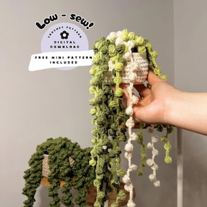 Puede incluir: Dos plantas de crochet en forma de collar de perlas, en verde y crema. Las plantas están en macetas pequeñas y tienen tallos largos y colgantes. El texto "Low-Sew! Crochet Pattern Digital Download Free Mini Pattern Included" está en un círculo morado con una flor blanca.