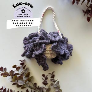 May include: A crocheted purple and white basket with a rope handle. The basket is on a white surface with green leaves in the background. The text "Low-Sew Crochet Pattern Digital Download Free Pattern Available on Instagram!" is on a purple and white circle.