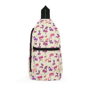 May include: A cream-coloured sling backpack featuring an all-over print of various pink and purple cartoon flamingos in different poses. The bag includes a black top handle, a black zip closure on the main compartment, and a front zipped pocket.