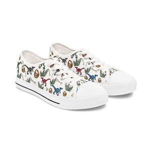 May include: A pair of white low-top canvas sneakers featuring a repeating pattern of colorful dinosaurs, fossilized eggs, green fern leaves, and scattered bones. The shoes have white laces, white rubber toe caps, and a thin black stripe along the sole.