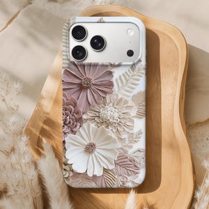 Könnte beinhalten: Eine weiße Handyhülle mit einem 3D-Blumendesign in Weiß-, Beige- und Mauve-Tönen. Die Hülle hat ein erhabenes Relief verschiedener Blumenarten und Blätter. Die Kameralinsen des Telefons sind oben sichtbar.