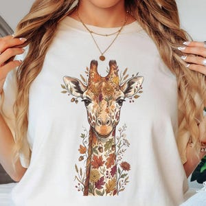 May include: An ivory t-shirt featuring a central graphic of a giraffe surrounded by autumn-themed foliage, including orange and brown leaves and small flowers. The giraffe is depicted in a realistic style with detailed fur patterns and warm tones.