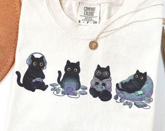 Puede incluir: Una camiseta de color marfil con un gráfico de cuatro gatos negros realizando diversas actividades de juego, como usar auriculares, utilizar un portátil, llevar gafas con sudadera y descansar en un puf con una consola portátil.