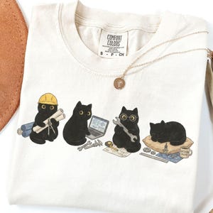 May include: An ivory t-shirt featuring a graphic of four black cats engaged in engineering tasks. One cat wears a yellow hard hat and holds blueprints, another uses a laptop, a third holds a spanner, and the fourth sleeps on a pillow.