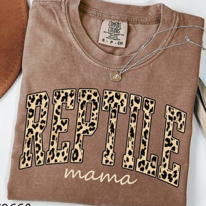 May include: A brown short-sleeved t-shirt with a crew neckline. The front features the word REPTILE in large block letters with a leopard print pattern, and the word mama in smaller white cursive script underneath. A silver necklace rests on top.