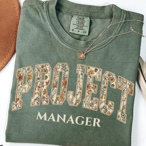 May include: A moss green short-sleeve t-shirt featuring the text PROJECT MANAGER in a collegiate font. The word PROJECT is filled with a floral pattern of orange, yellow, and brown leaves and flowers. The shirt has a crew neck and a size S label.