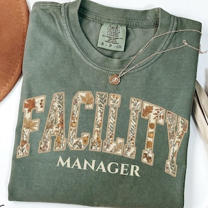 May include: A moss green crewneck t-shirt featuring the text FACILITY MANAGER in a collegiate font. The letters are filled with a botanical pattern of autumn leaves, acorns, and flowers. The shirt has a Comfort Colors label in the neck.
