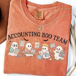 May include: A terracotta-colored t-shirt featuring a graphic of four ghosts working in an office setting. The text "ACCOUNTING BOO TEAM" is printed above the ghosts, who are surrounded by office supplies, documents, and small Halloween icons.
