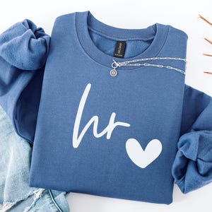 May include: A blue crewneck sweatshirt featuring the white text "hr" in a cursive script followed by a white heart graphic. The garment is displayed alongside light blue distressed denim jeans and a silver chain necklace.