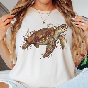 May include: An ivory t-shirt featuring a graphic illustration of a sea turtle adorned with autumn-colored leaves, small flowers, and acorns. The turtle is depicted in shades of brown and green, centered on the chest of the short-sleeved garment.