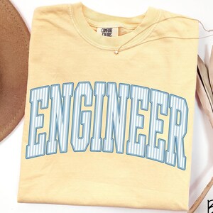May include: A butter yellow short-sleeve t-shirt with a crew neckline and a Comfort Colors brand label. The front features the word ENGINEER in large, arched, block letters with a light blue and white vertical pinstripe pattern.