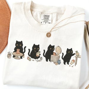 May include: An ivory t-shirt featuring a graphic of four black cats engaged in pottery making. The illustration shows cats using a pottery wheel, painting a vase, using a kiln, and placing a finished vase on a small wooden table. The shirt has a Comfort Colors brand label at the neck.