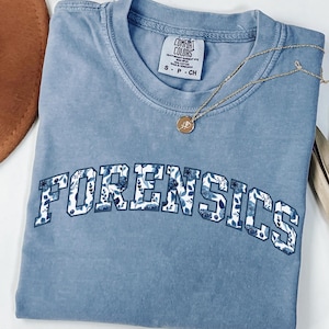 Puede incluir: Una camiseta azul con la palabra "FORENSICS" en un diseño floral en la parte delantera. Un collar dorado con un colgante descansa sobre la camisa. La camisa está doblada y colocada sobre una superficie blanca.