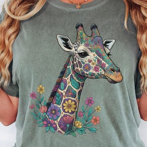 May include: A moss green short-sleeve t-shirt featuring a graphic print of a giraffe head and neck. The giraffe is decorated with colourful floral patterns in shades of purple, yellow, pink, and blue, surrounded by small flowers at the base.