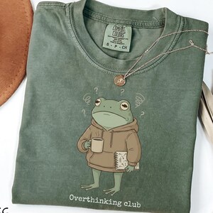 May include: Moss green t-shirt featuring a cartoon frog in a hoodie, holding a mug and notepad, with a perplexed expression and question marks. The text "Overthinking club" is printed below the frog. A gold necklace is also visible.