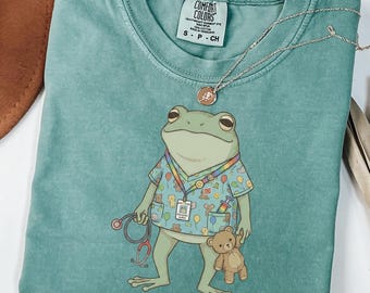 May include: Light green t-shirt with a frog graphic in a pediatric nurse's uniform, holding a stethoscope and a teddy bear. The text "Pediatric care is my swamp" is printed at the bottom. A silver necklace is draped across the shirt.