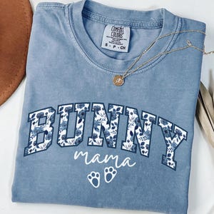 May include: A blue Comfort Colors t-shirt with the word "BUNNY" in a floral design and the word "mama" in script. Below are two white bunny paw prints. A gold necklace with a pendant is on the shirt.
