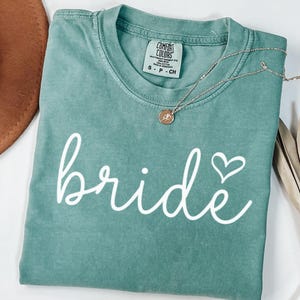 May include: A teal t-shirt with the word "bride" in white script and a heart symbol. A gold necklace with a pendant is draped across the shirt. The shirt is folded, and a brown hat is visible in the background. A dried plant is on the right.