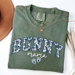 Puede incluir: Una camiseta de manga corta color verde oliva con la palabra BUNNY en letras de bloque rellenas con un patr&oacute;n floral azul, la palabra mama en letra cursiva blanca y dos huellas de pata blancas. La camiseta tiene una etiqueta Comfort Colors.