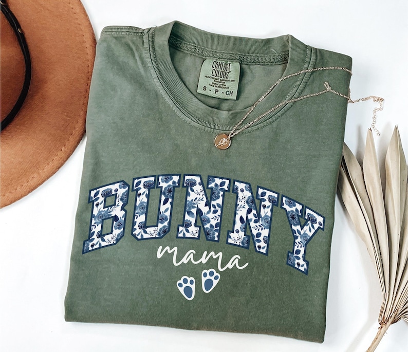 Puede incluir: Una camiseta de manga corta color verde oliva con la palabra BUNNY en letras de bloque rellenas con un patr&oacute;n floral azul, la palabra mama en letra cursiva blanca y dos huellas de pata blancas. La camiseta tiene una etiqueta Comfort Colors.
