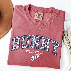 Puede incluir: Una camiseta de cuello redondo color rosa empolvado con la palabra BUNNY en letras de bloque rellenas de un estampado floral azul. Debajo, el texto mama est&aacute; impreso en letra cursiva blanca, seguido de dos huellas de conejo blancas. Talla S.