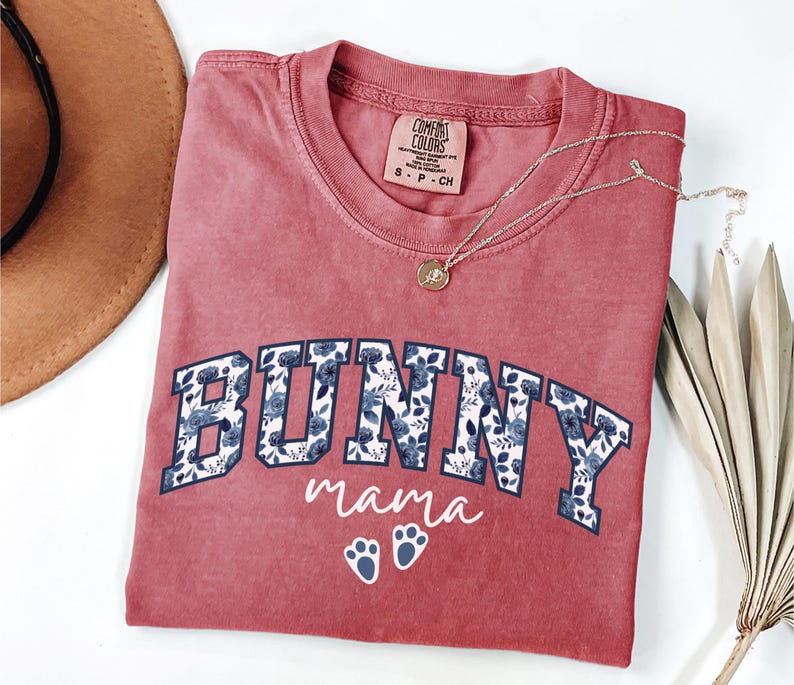 Puede incluir: Una camiseta de cuello redondo color rosa empolvado con la palabra BUNNY en letras de bloque rellenas de un estampado floral azul. Debajo, el texto mama est&aacute; impreso en letra cursiva blanca, seguido de dos huellas de conejo blancas. Talla S.