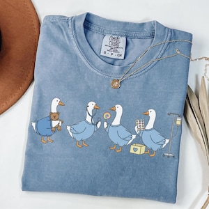 May include: A blue Comfort Colors t-shirt featuring a graphic of four geese dressed as medical professionals. One goose holds a teddy bear, another a stethoscope, a third a lollipop, and the last, pills and an IV drip. A gold necklace rests on the shirt.