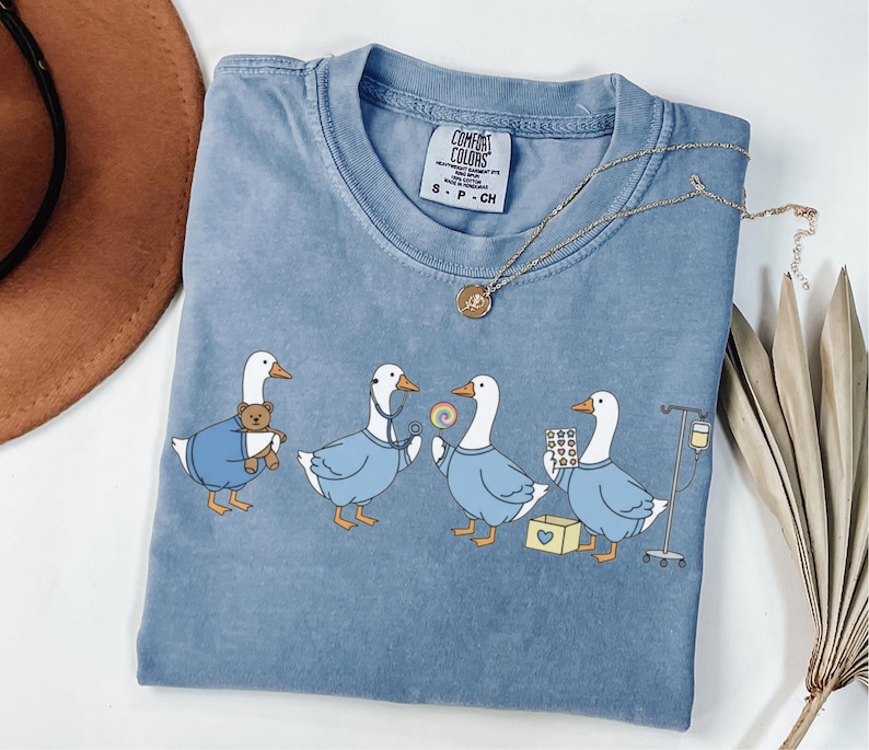May include: A blue Comfort Colors t-shirt featuring a graphic of four geese dressed as medical professionals. One goose holds a teddy bear, another a stethoscope, a third a lollipop, and the last, pills and an IV drip. A gold necklace rests on the shirt.