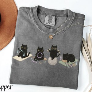 Puede incluir: Una camiseta de cuello redondo en color gris carb&oacute;n con un gr&aacute;fico de cuatro gatos negros realizando diversos ejercicios, incluyendo el uso de una banda de resistencia, un aro de pilates, mancuernas sobre una pelota de yoga y descanso.