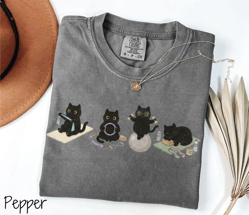 Puede incluir: Una camiseta de cuello redondo en color gris carb&oacute;n con un gr&aacute;fico de cuatro gatos negros realizando diversos ejercicios, incluyendo el uso de una banda de resistencia, un aro de pilates, mancuernas sobre una pelota de yoga y descanso.