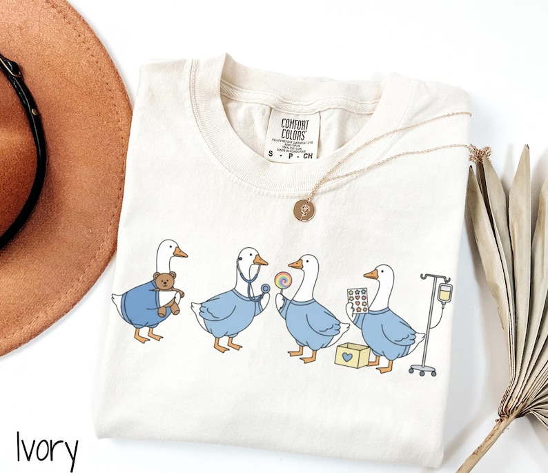 May include: Ivory-colored t-shirt featuring a line of cartoon geese dressed as medical professionals. Each goose is wearing a blue uniform and holding medical items like a teddy bear, stethoscope, lollipop, pills, and IV drip. A gold necklace is draped over the shirt.