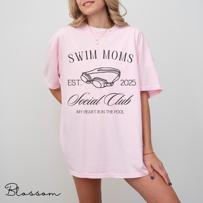 Puede incluir: Una camiseta rosa claro con texto negro que dice SWIM MOMS EST. 2025 Social Club MY HEART IS IN THE POOL, junto a un dibujo lineal central de unas gafas de nataci&oacute;n. La camiseta tiene mangas cortas y cuello redondo.