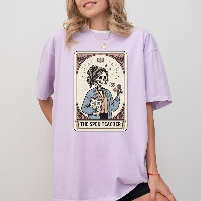 Puede incluir: Una camiseta de color lila claro con un dise&ntilde;o de carta del tarot. El gr&aacute;fico representa a un profesor esqueleto sosteniendo una carpeta y objetos coloridos, con el texto "THE SPED TEACHER" en la parte inferior. La camiseta tiene un corte holgado y extragrande.