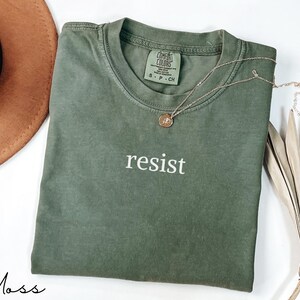 Puede incluir: Una camiseta verde musgo con la palabra "resist" impresa en blanco. Un collar dorado con un colgante est&aacute; drapeado sobre la camiseta. La camiseta est&aacute; doblada sobre una superficie blanca, con un sombrero marr&oacute;n y hojas de palma secas en el fondo.