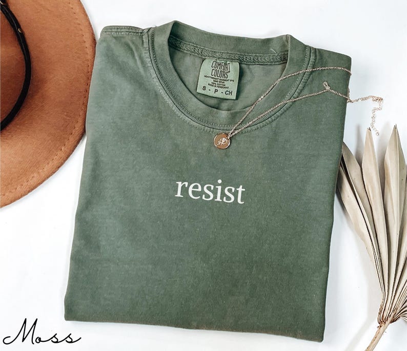 Puede incluir: Una camiseta verde musgo con la palabra "resist" impresa en blanco. Un collar dorado con un colgante est&aacute; drapeado sobre la camiseta. La camiseta est&aacute; doblada sobre una superficie blanca, con un sombrero marr&oacute;n y hojas de palma secas en el fondo.