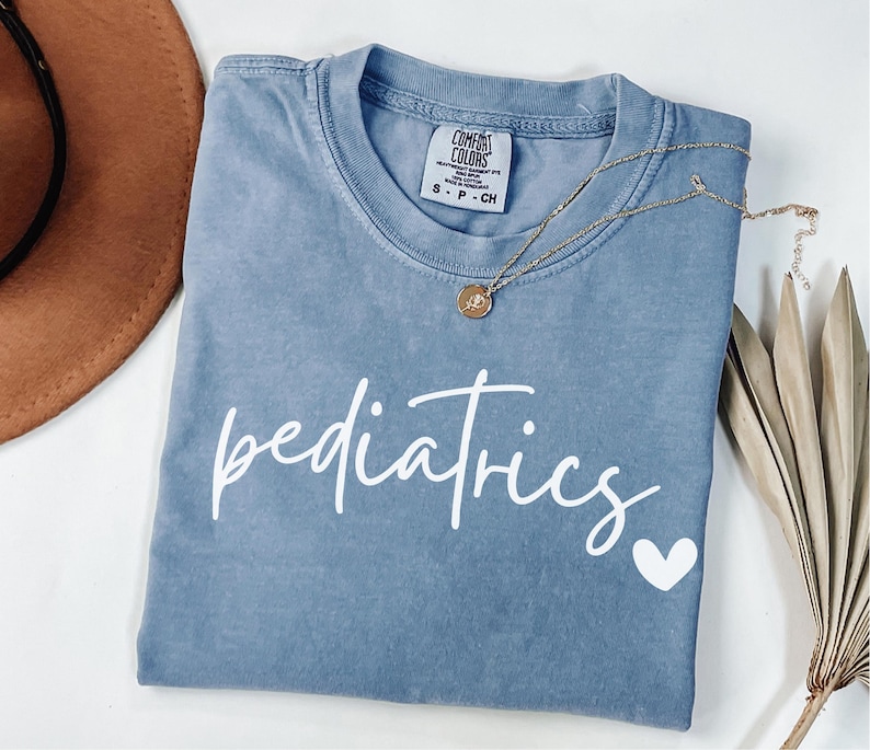 May include: A blue Comfort Colors t-shirt with the word "pediatrics" in white script and a white heart. A gold necklace with a pendant is draped across the shirt. A brown hat is visible in the upper left corner.