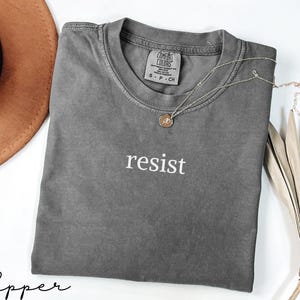 Puede incluir: Una camiseta gris Comfort Colors con la palabra "resist" en letras blancas. Un collar plateado con un peque&ntilde;o colgante descansa sobre la camiseta. Un sombrero marr&oacute;n y una planta seca est&aacute;n en el fondo.