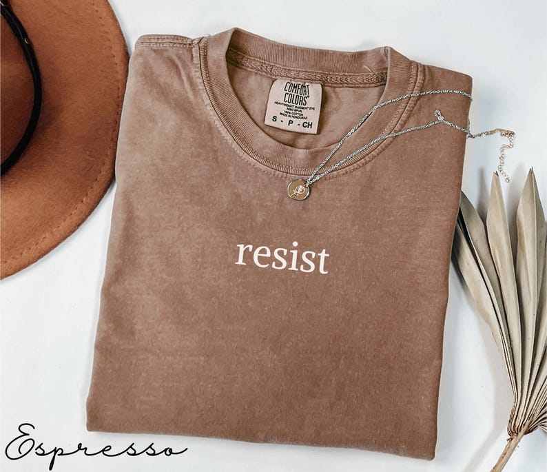 Puede incluir: Una camiseta marr&oacute;n de manga corta con la palabra "resist" impresa en blanco. La camiseta tiene cuello redondo y una peque&ntilde;a etiqueta en el cuello. Un collar plateado con un colgante est&aacute; drapeado sobre la camiseta. La palabra "Espresso" est&aacute; escrita en cursiva en la parte inferior.